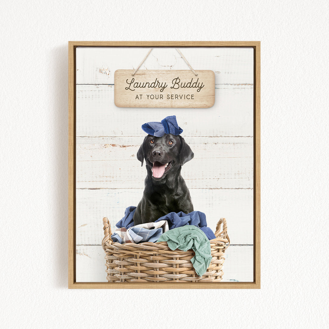 A black dog wearing a blue hat sits in a wicker basket filled with clothes, with a wooden sign above that reads "Laundry Buddy at your service".