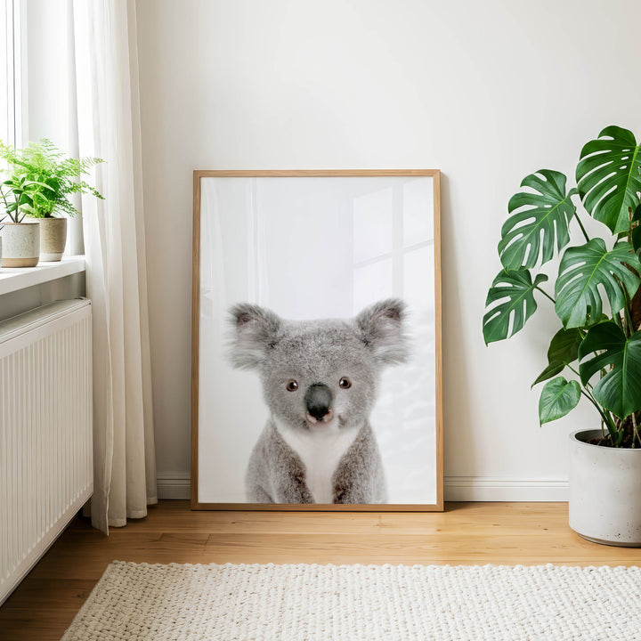 a large framed picture of a koala bear, which is the central focus of the room. The koala bear is sitting in front of a window, and there are two potted plants in the room, one on the left side and the other on the right side. The room has a white wall and a wooden floor, creating a cozy and natural atmosphere.