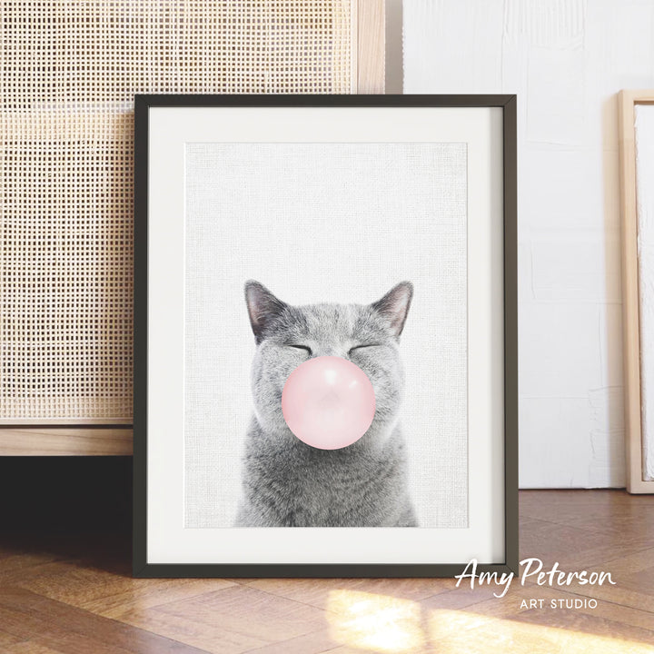 A framed image of a gray cat blowing a pink bubble gum bubble.