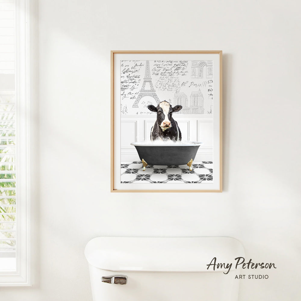 A framed print of a cow sitting in a bathtub is hanging on a white wall, with a window and a toilet visible in the background.