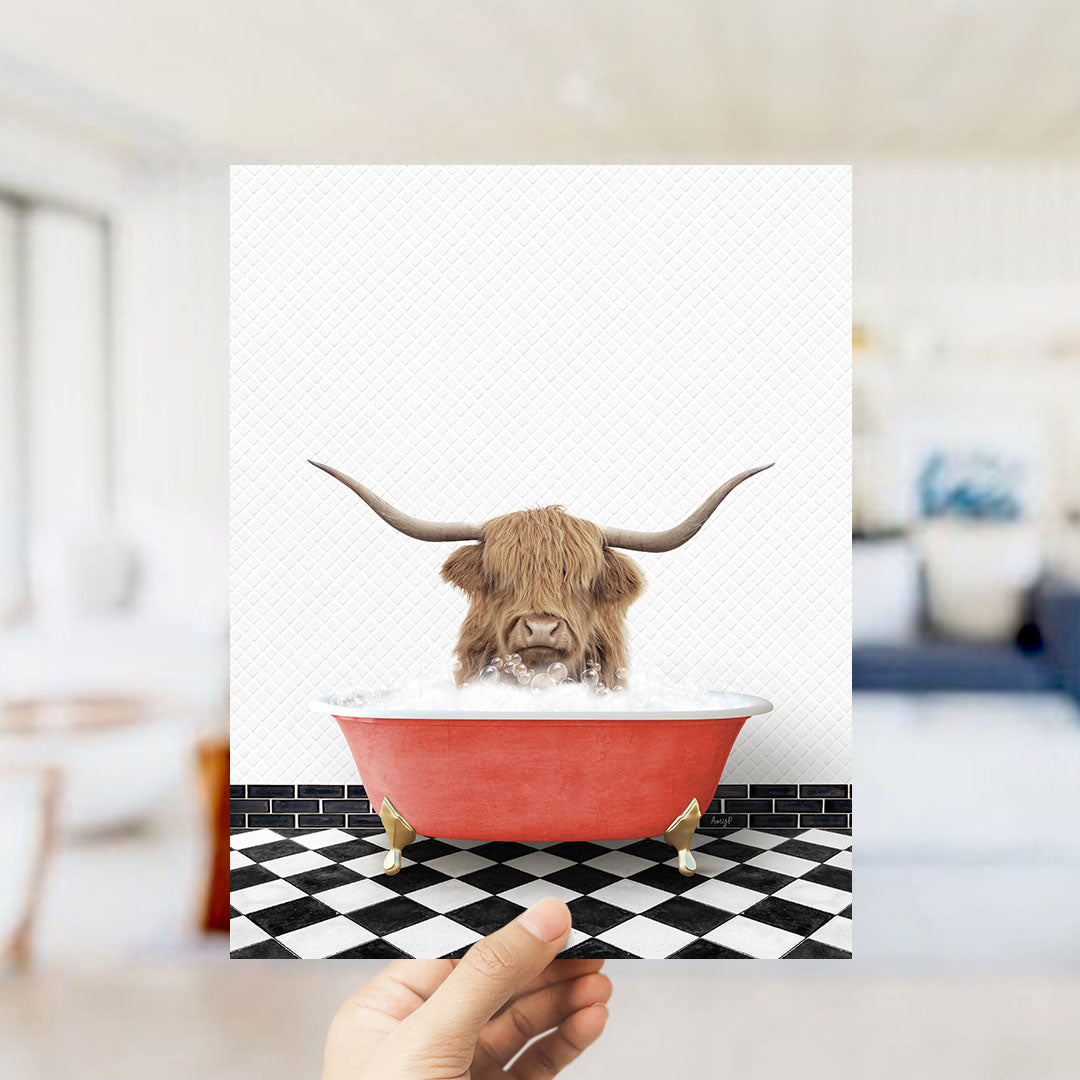 A hand holds up a framed image depicting a Highland cow with large horns sitting in a red bathtub on a black and white checkered floor.