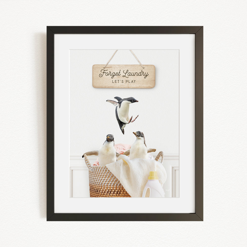 A framed image of three penguins in a laundry basket, with a wooden sign above them that says "Forget Laundry, Let's Play".