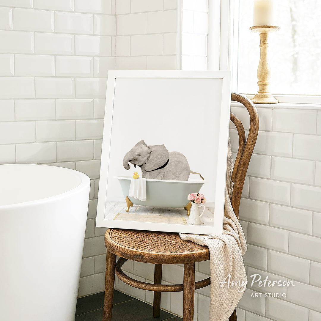 A framed image of a gray elephant sitting in a bathtub, with a vase of flowers and a towel nearby, is displayed on a wooden chair in a bathroom setting.