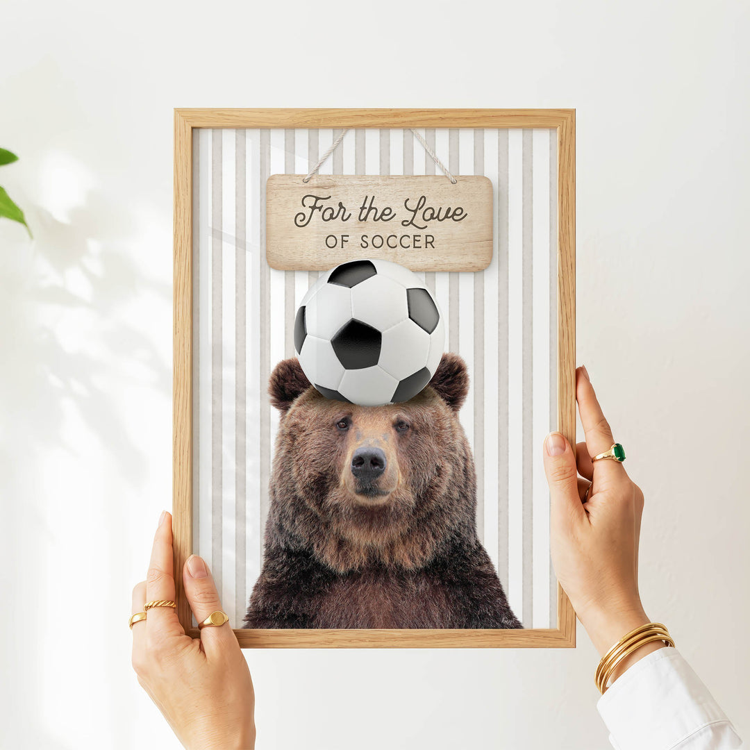 A person's hands are holding a framed picture of a bear with a soccer ball on its head, with a sign above the bear that says "For the Love of Soccer".