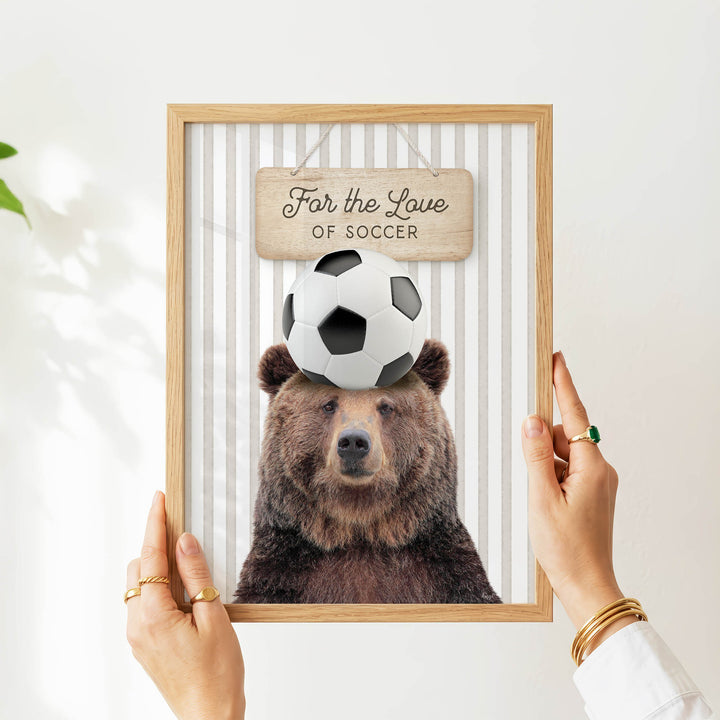 A person's hands are holding a framed picture of a bear with a soccer ball on its head, with a sign above the bear that says "For the Love of Soccer".