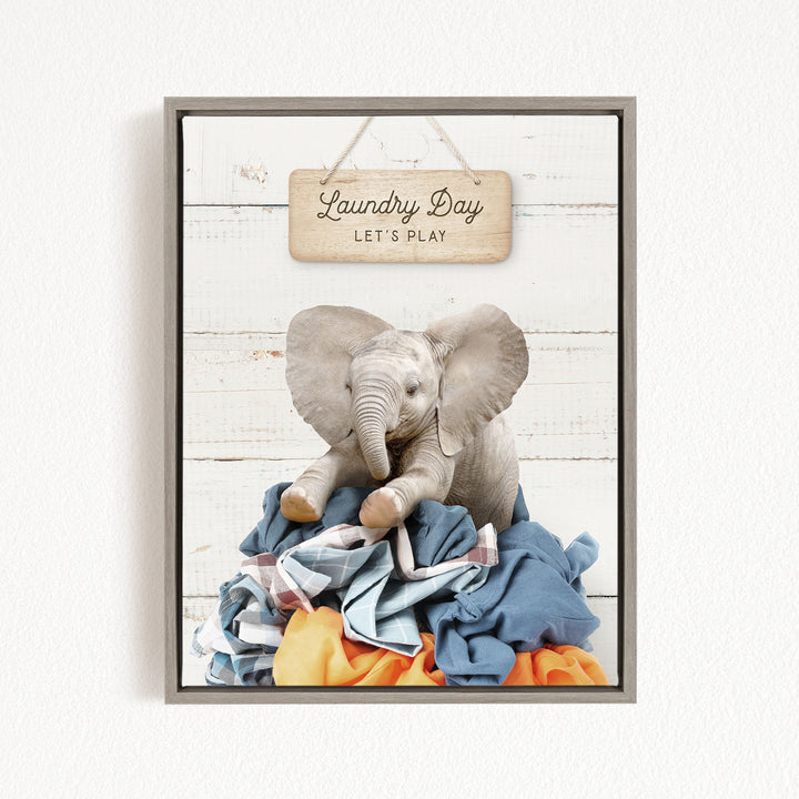 A framed image of a gray elephant sitting atop a pile of laundry, with a wooden sign above that reads "Laundry Day Let's Play".