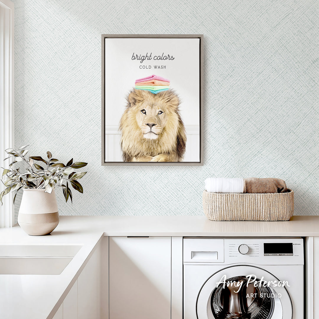a framed poster of a lion with a colorful hat on its head, hanging on a wall above a white countertop with a potted plant and a basket of towels.