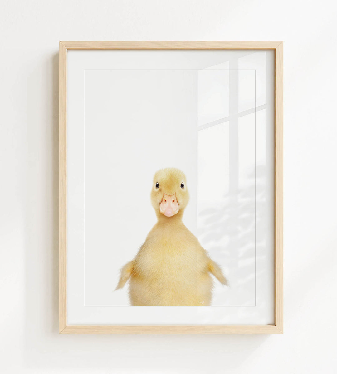 A cute yellow duck with a pink beak is the main subject of the framed print, which is hung on a white wall.