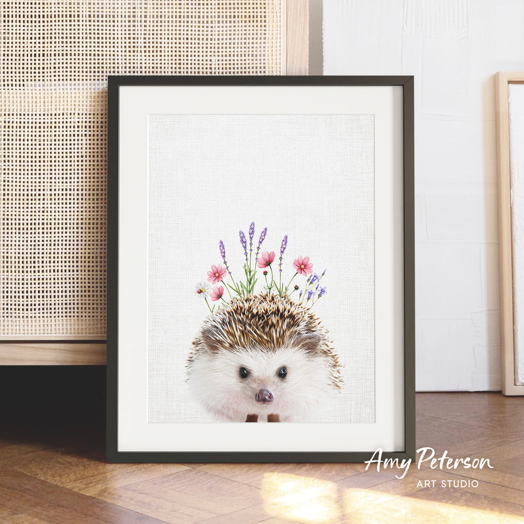 A framed print of a hedgehog with flowers on its head, placed on a wooden floor against a white wall.