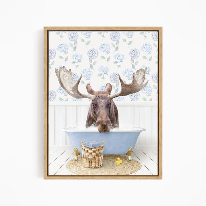 A moose with large antlers is sitting in a blue bathtub filled with water, surrounded by a wicker basket and rubber ducks.