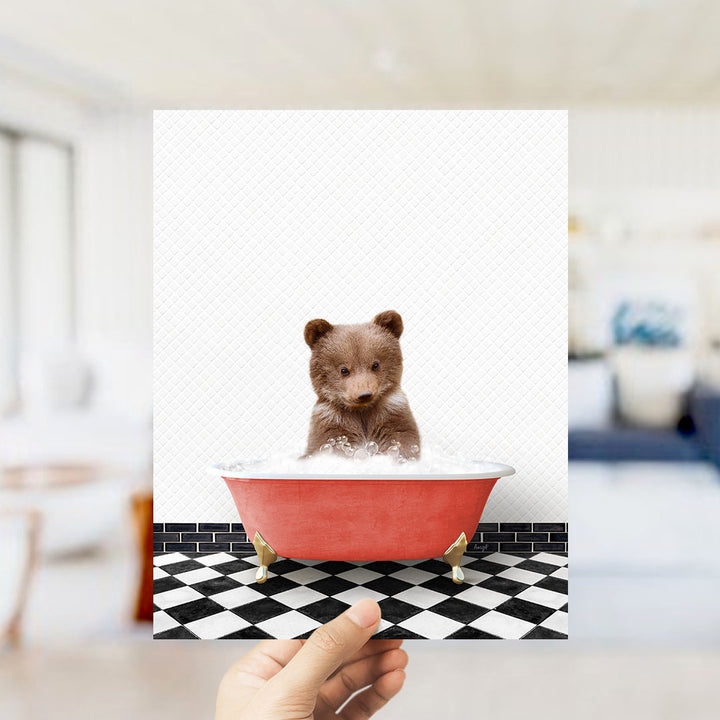 A hand is holding up a framed image of a small brown bear sitting in a red bathtub filled with bubbles, set against a black and white checkered floor.