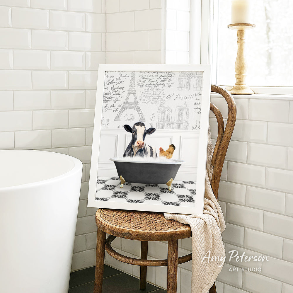 A framed image of a cow sitting in a bathtub, with a chicken nearby, is displayed on a wooden chair in a bathroom setting.