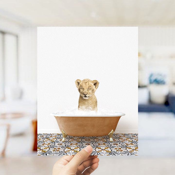 A hand holds up a framed image of a young lion cub sitting in a bathtub filled with water, surrounded by a patterned tile floor.