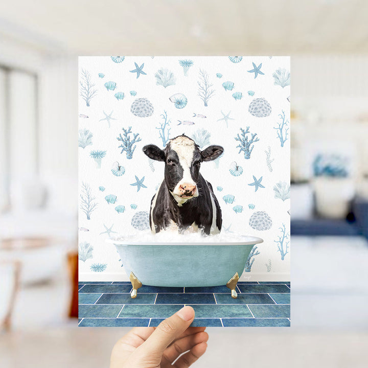 A hand holds up a printed image of a cow sitting in a bathtub filled with bubbles, surrounded by a pattern of seashells, starfish, and other marine life.