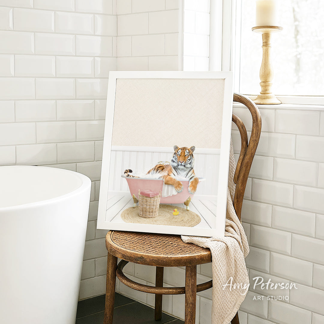 A framed image of a tiger sitting in a pink bathtub, surrounded by a basket of toys, is displayed on a wooden chair in a bathroom setting.