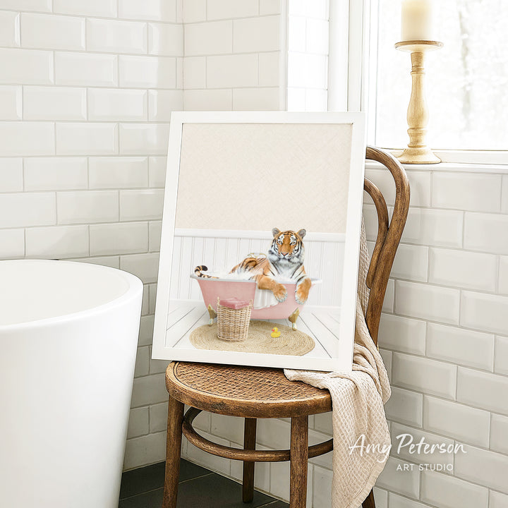 A framed image of a tiger sitting in a pink bathtub, surrounded by a basket of toys, is displayed on a wooden chair in a bathroom setting.