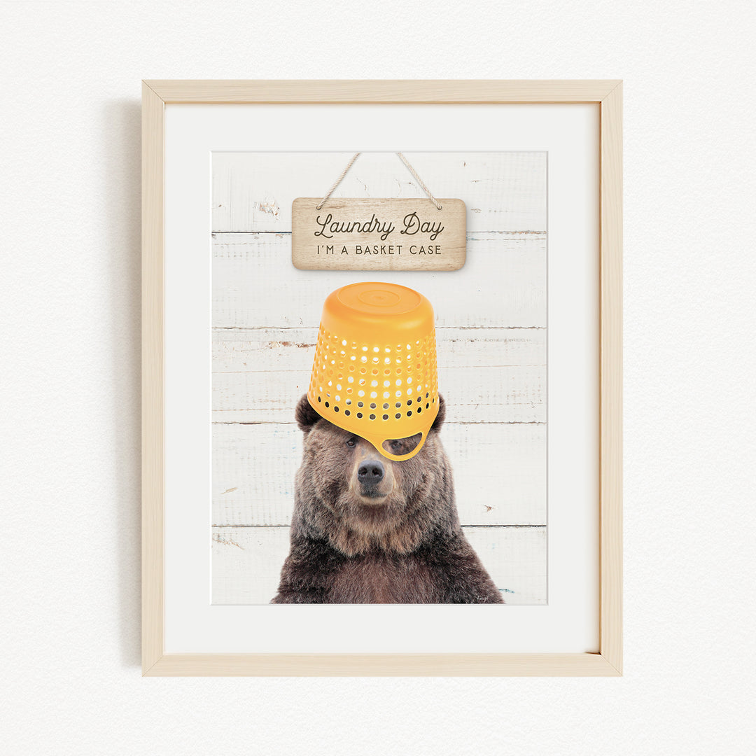 A framed image of a bear wearing a yellow basket on its head, with a sign above that says "Laundry Day I'm a basket case".