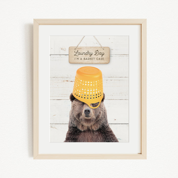 A framed image of a bear wearing a yellow basket on its head, with a sign above that says "Laundry Day I'm a basket case".