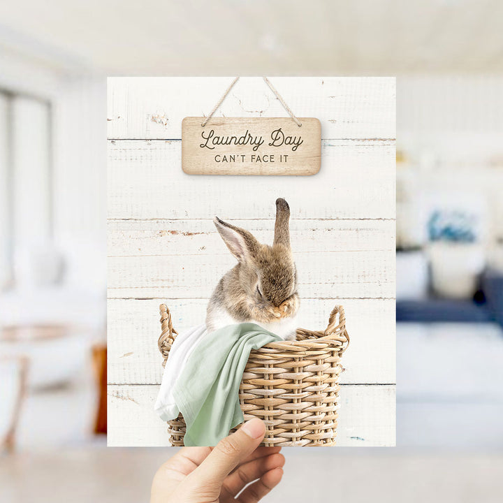 A hand holding a card with a picture of a rabbit sitting in a wicker basket, with a sign above that says "Laundry Day Can't Face It".