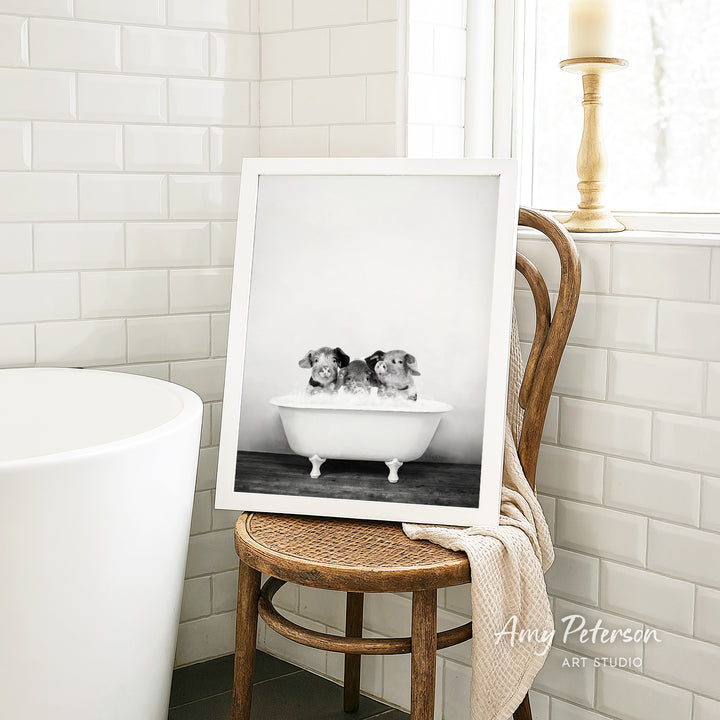 A framed black and white photograph of three puppies sitting in a bathtub, with a wooden chair and a white tiled wall in the background.