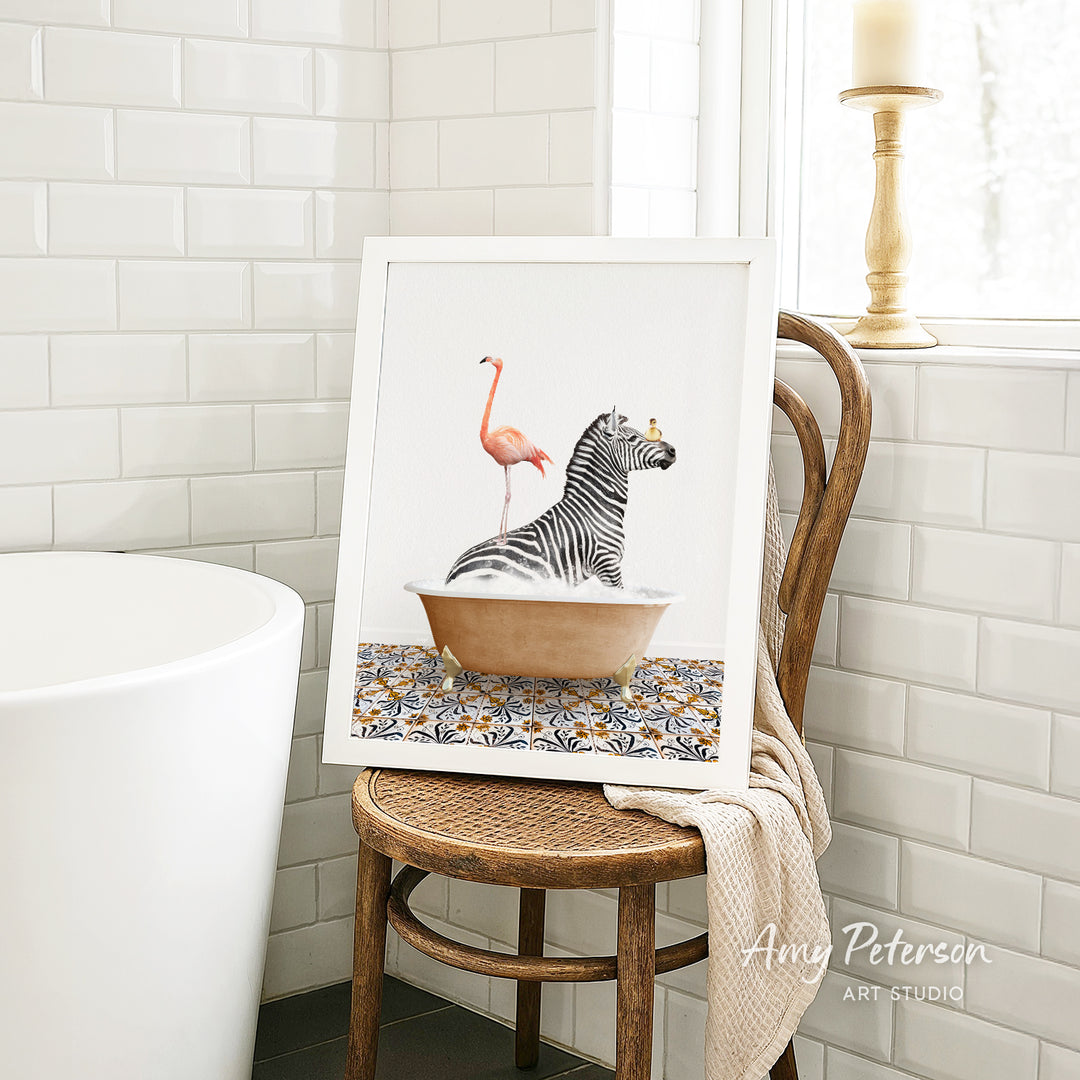 A framed image of a zebra sitting in a bathtub, with a pink flamingo standing on its back, is displayed on a wooden chair in a bathroom setting.