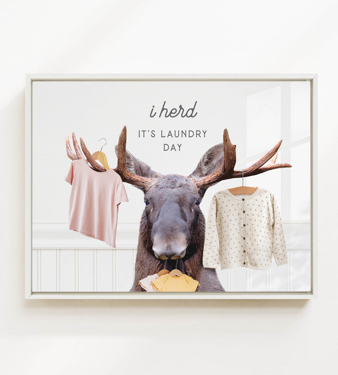 A framed image depicting a moose with antlers holding clothes, accompanied by the text "i herd, it's laundry day".