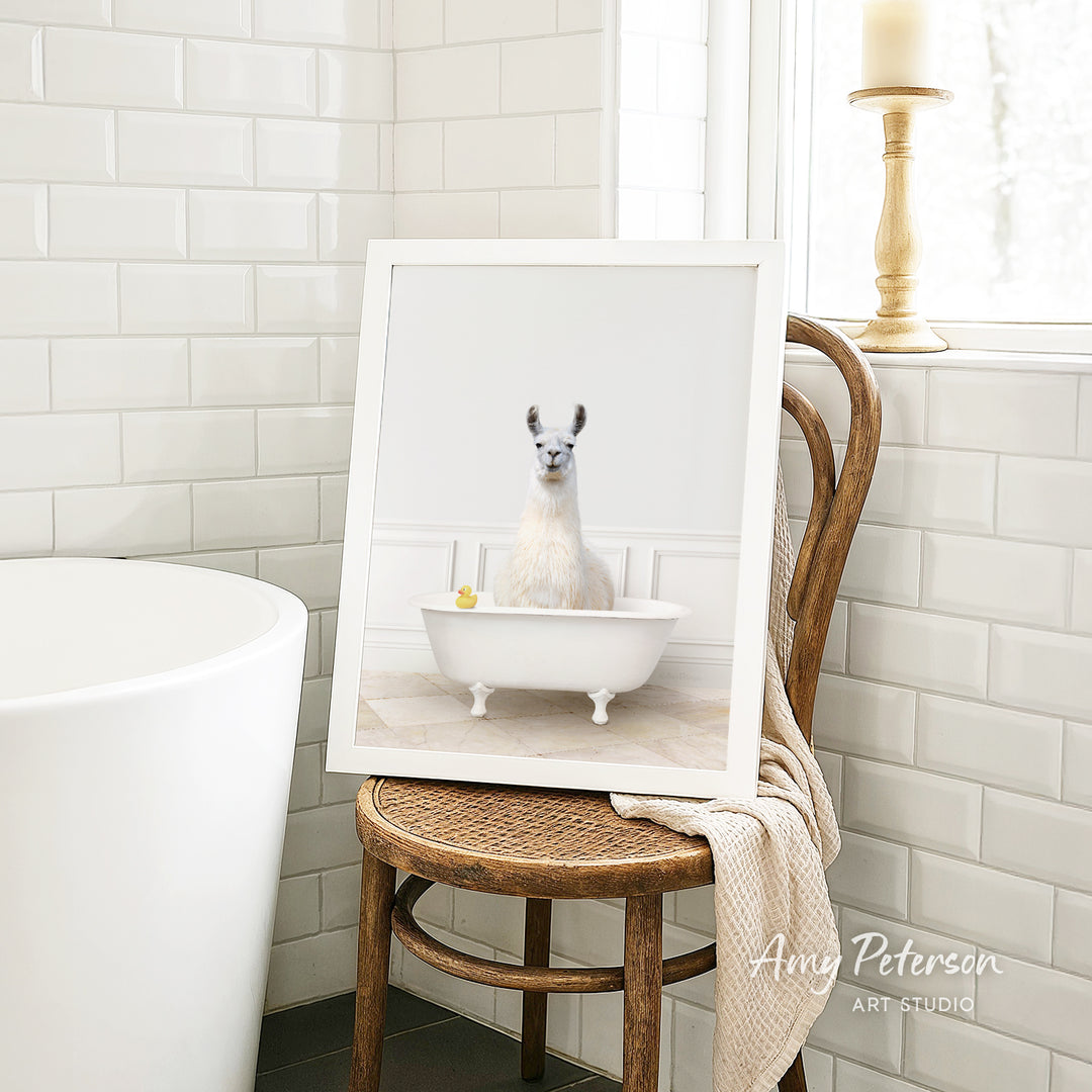 A framed image of a white llama sitting in a white bathtub is displayed on a wooden chair in a bathroom setting.