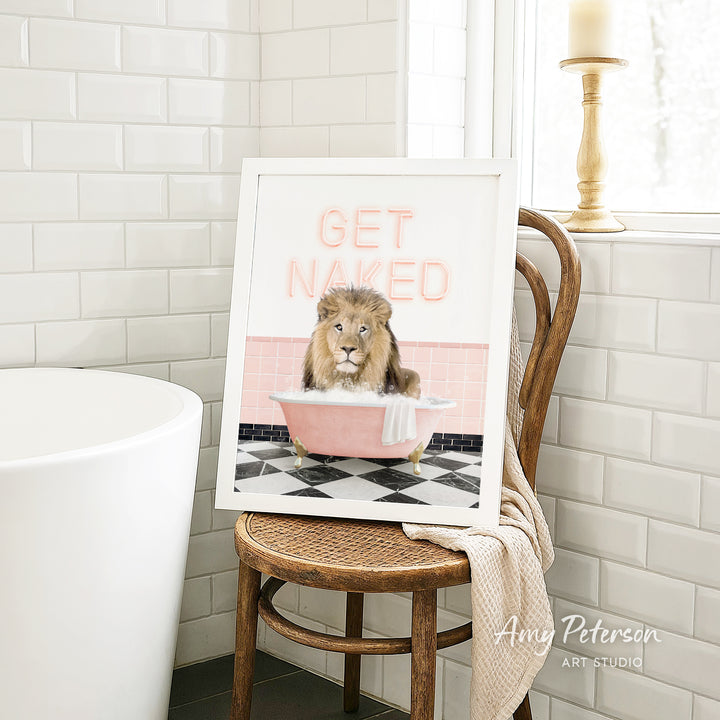 A framed image of a lion sitting in a pink bathtub with the text "GET NAUGED" on it, placed on a wooden chair in a bathroom setting.
