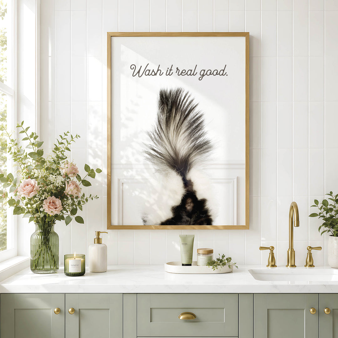 a bathroom counter with a framed poster of a skunk's tail, a vase of pink flowers, and various toiletries such as a soap dispenser and a candle.