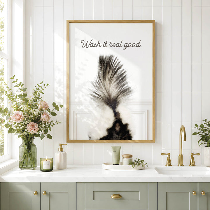 a bathroom counter with a framed poster of a skunk's tail, a vase of pink flowers, and various toiletries such as a soap dispenser and a candle.