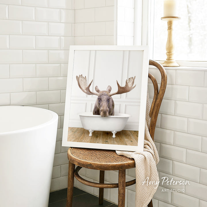 A framed image of a moose sitting in a bathtub is displayed on a wooden chair in a bathroom setting.