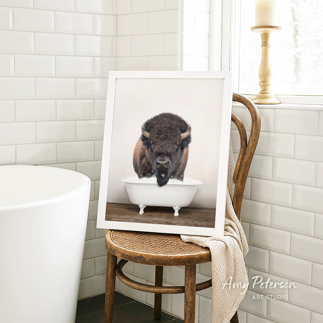 A framed photograph of a bison resting in a white bathtub is displayed on a wooden chair in a bathroom setting.