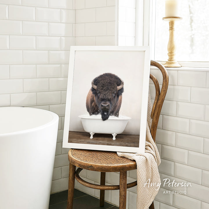 A framed photograph of a bison resting in a white bathtub is displayed on a wooden chair in a bathroom setting.