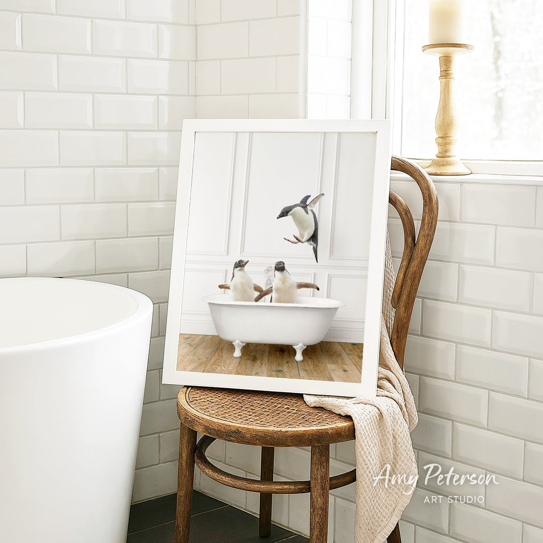 A framed image of two penguins sitting in a bathtub, with one penguin standing and the other sitting, is displayed on a wooden chair in a bathroom setting.