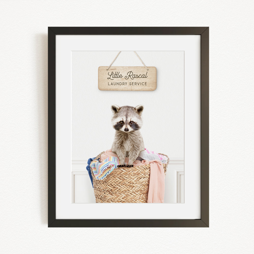 A raccoon sitting in a woven laundry basket, with a sign above that reads "Little Racal Laundry Service".
