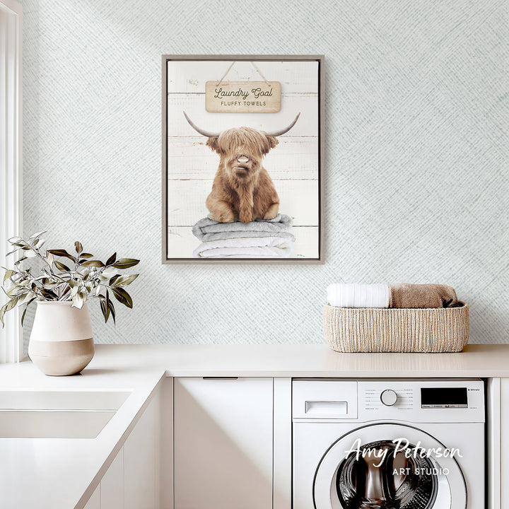 a framed picture of a Highland cow with a sign that says "Laundry Bowl Fluffy Towel" above it, hanging on a wall above a white countertop with a basket and a potted plant.
