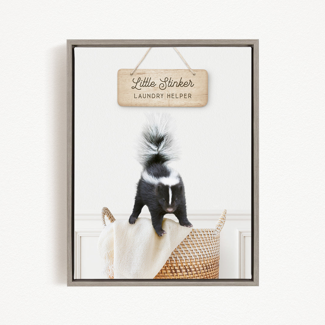 A framed image of a black and white skunk standing on a woven basket, with a wooden sign above it that reads "Little Stinker Laundry Helper".
