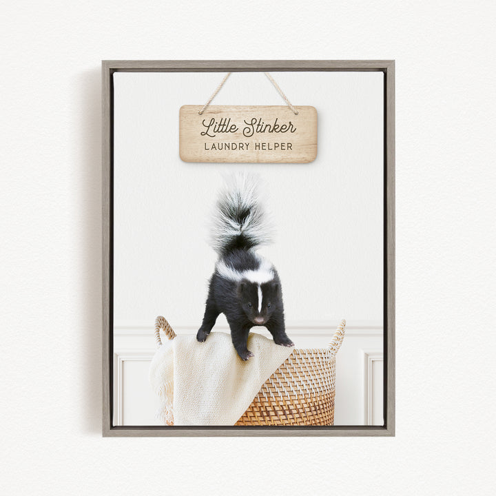 A framed image of a black and white skunk standing on a woven basket, with a wooden sign above it that reads "Little Stinker Laundry Helper".