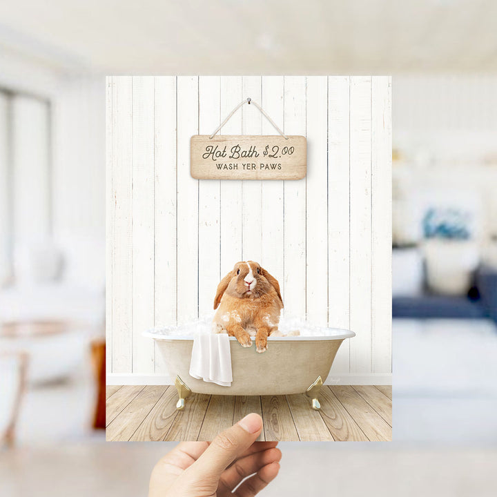 A hand holding a framed image of a brown rabbit sitting in a bathtub, with a sign above it that says "Hot Bath $2.00 WASH YOUR Paws".