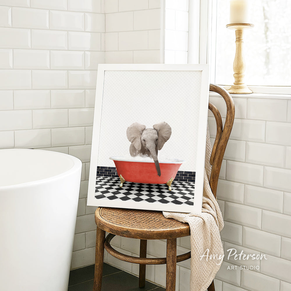 A framed image of a baby elephant sitting in a red bathtub, surrounded by a black and white checkered floor, is displayed on a wooden chair in a bathroom setting.