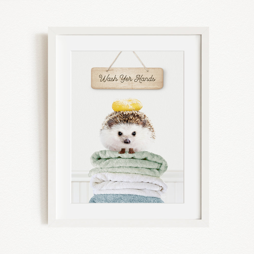 A framed image of a hedgehog sitting atop a stack of towels, with a wooden sign above that reads "Wash Your Hands".