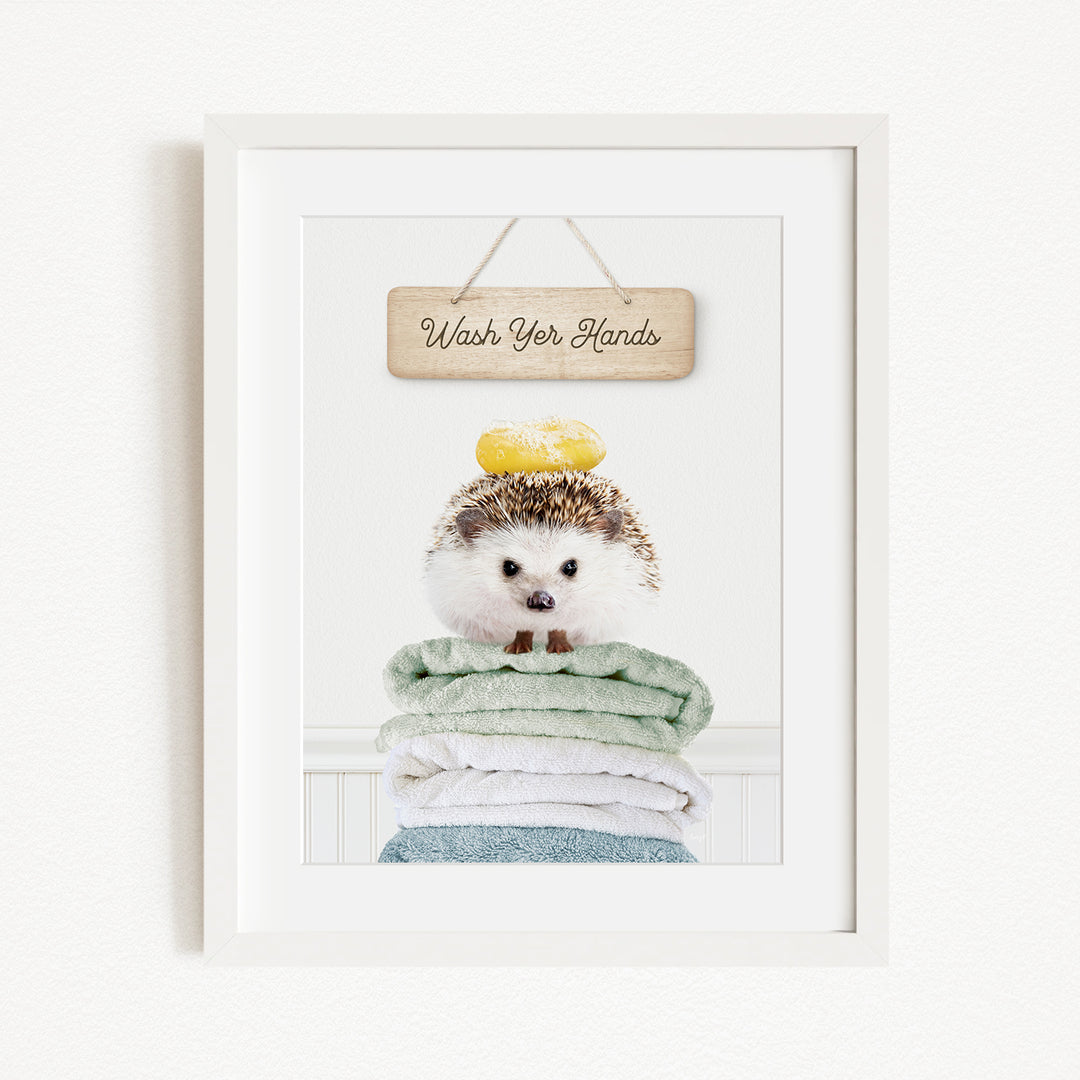 A framed image of a hedgehog sitting atop a stack of towels, with a wooden sign above that reads "Wash Your Hands".