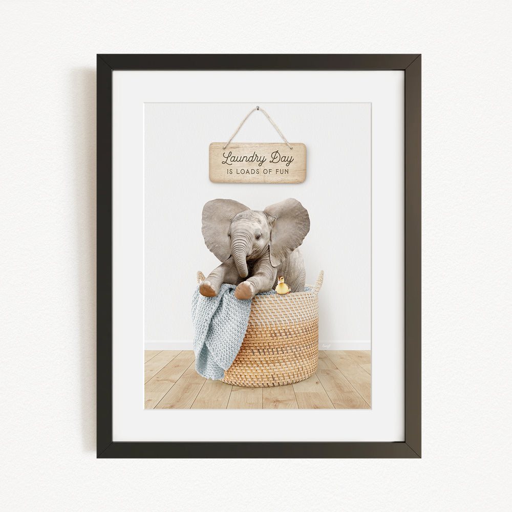 A framed image of a baby elephant sitting in a woven basket, with a wooden sign above that reads "Laundry Day is loads of fun".