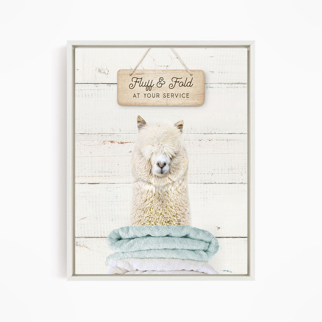 A fluffy alpaca sitting on a stack of blue towels, with a wooden sign above that says "Fuffy & Fold at your service".