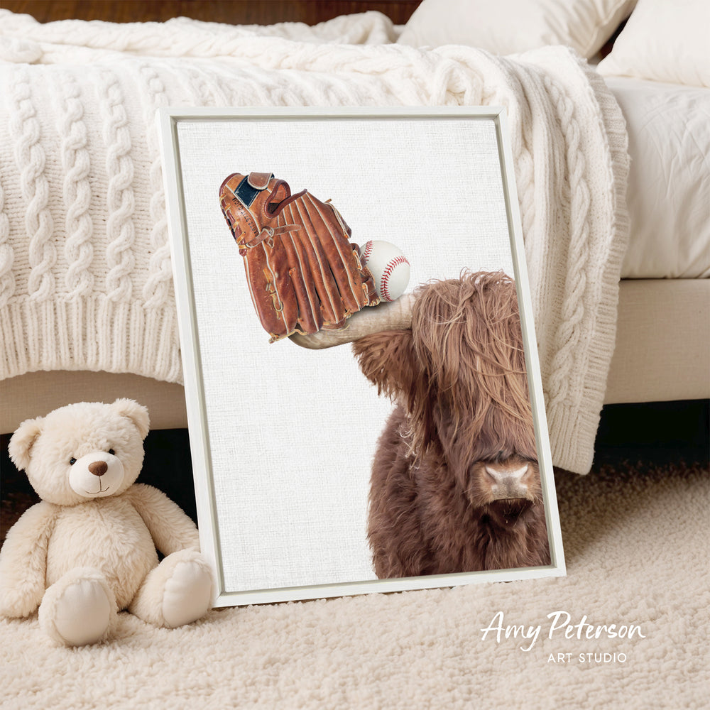 A framed print of a teddy bear and a baseball glove is displayed on a white bedspread, with a stuffed animal to the left.