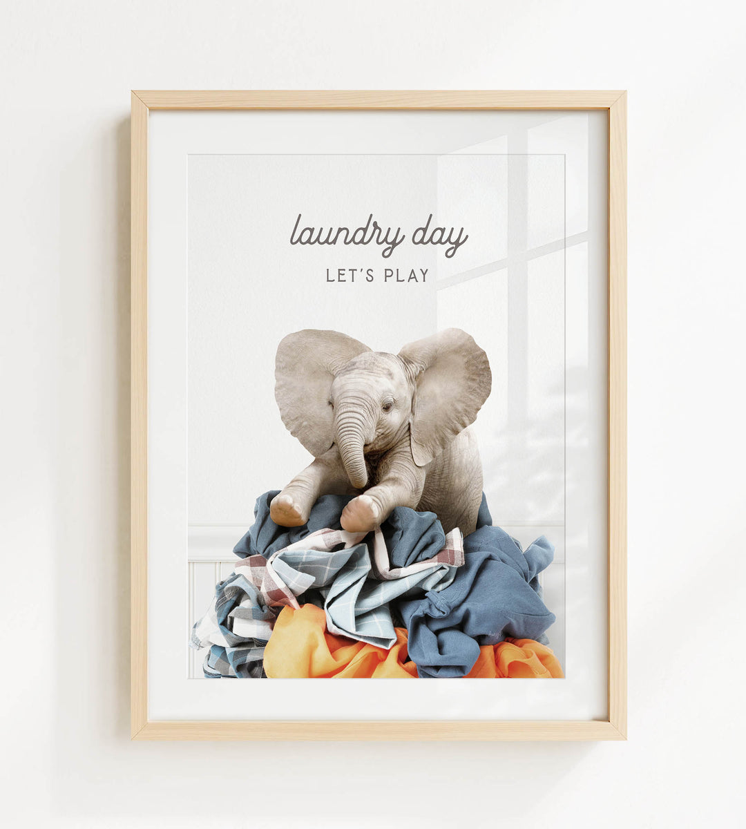 A framed image of a baby elephant sitting atop a pile of laundry, with the text "laundry day let's play" written above it.
