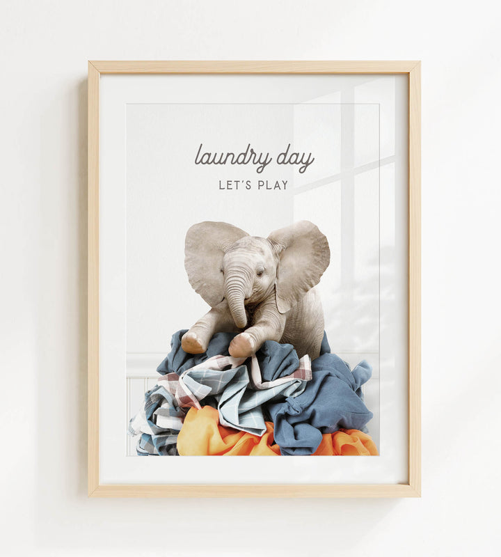 A framed image of a baby elephant sitting atop a pile of laundry, with the text "laundry day let's play" written above it.