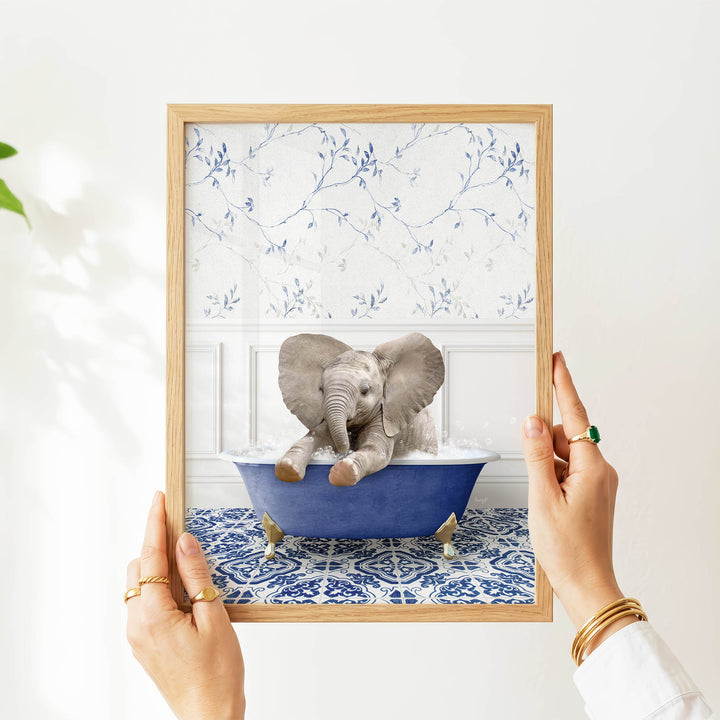 A person's hands are holding a framed picture of a baby elephant sitting in a blue bathtub, with a patterned wall behind it.