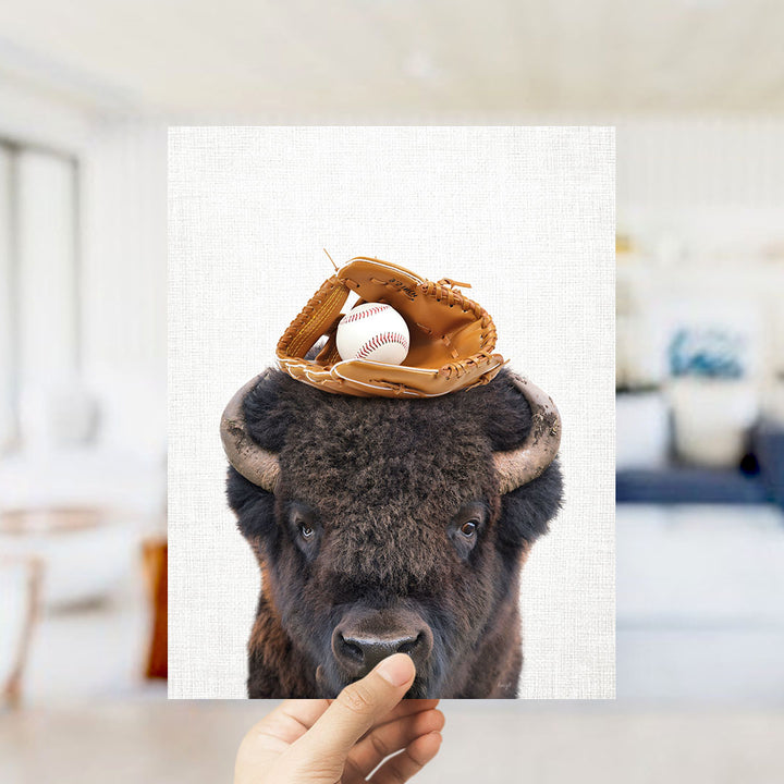 A hand holds up a framed image of a buffalo with a baseball glove on its head.