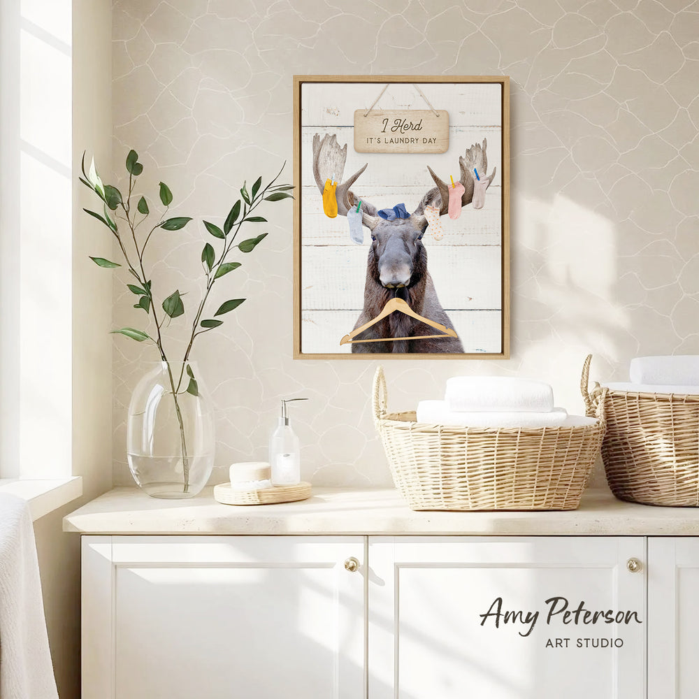 a framed picture of a moose with a sign that says "I herd it's laundry day" hanging on a wall above a white cabinet.